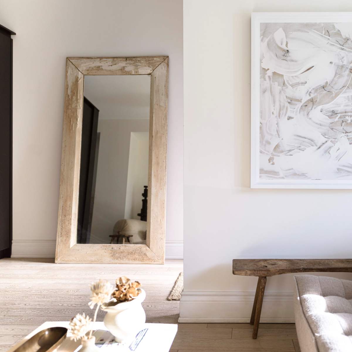 A large rustic mirror stands against the wall next to a simple wooden bench and a piece of abstract artwork hanging above it.