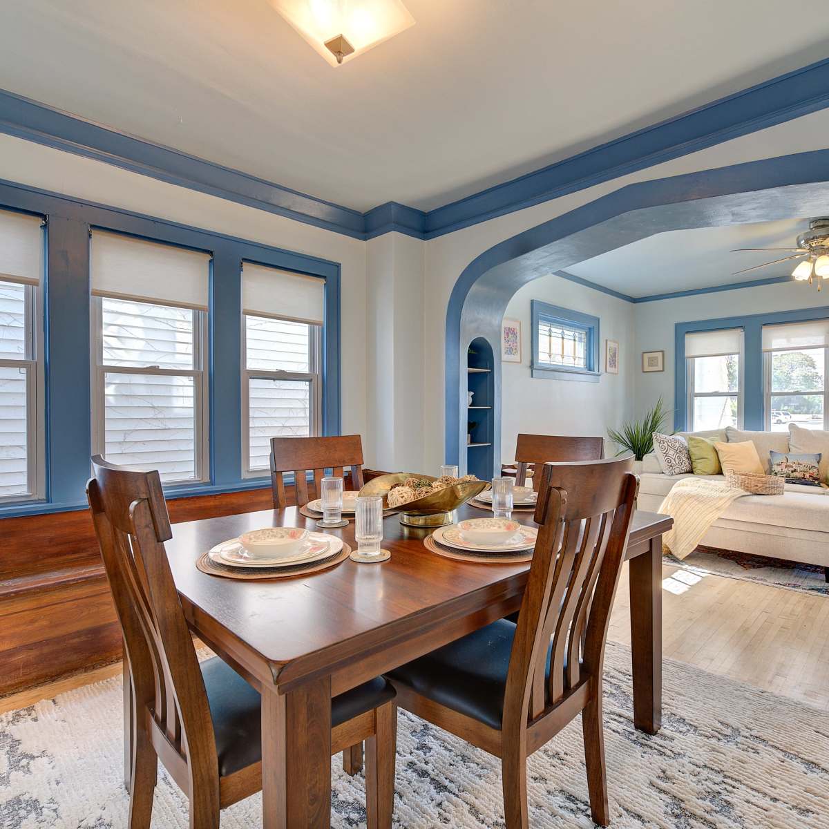 A dining area features a wooden table set for four, surrounded by chairs, with an archway leading to a cozy living space filled with natural light.