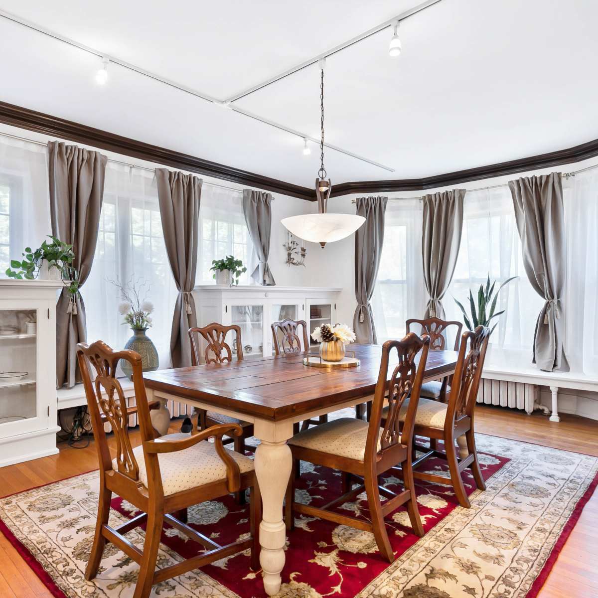 A wooden dining table surrounded by chairs is set in a well-lit room featuring bay windows, white cabinetry, and draped curtains.