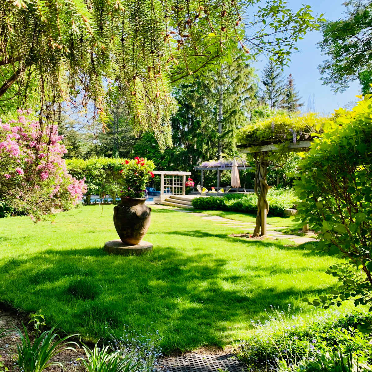 A landscaped garden features a circular lawn surrounded by flowering shrubs and a large decorative pot at the center.