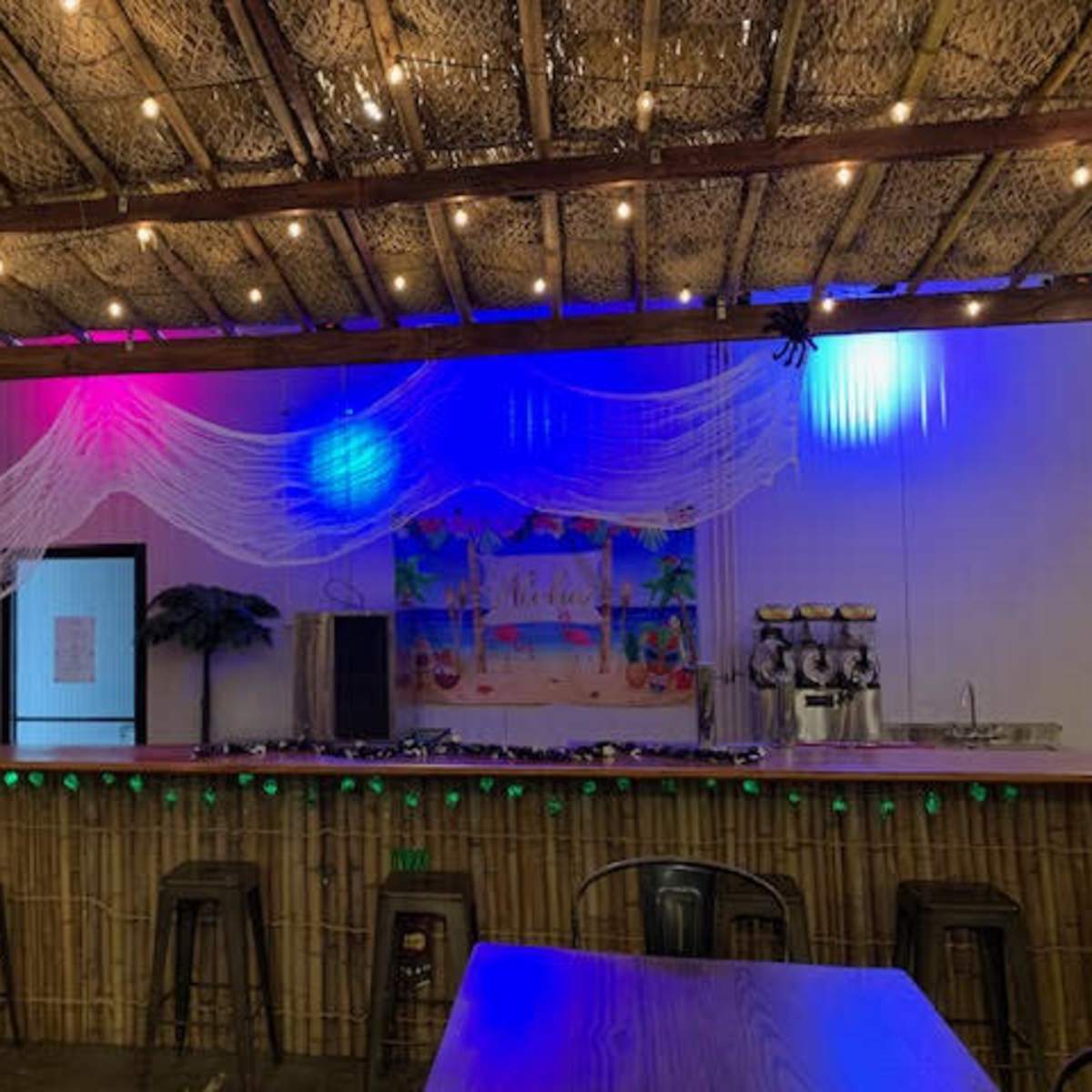 The interior of a bar features a wooden counter, bamboo decor, and colorful lights draped across the ceiling.