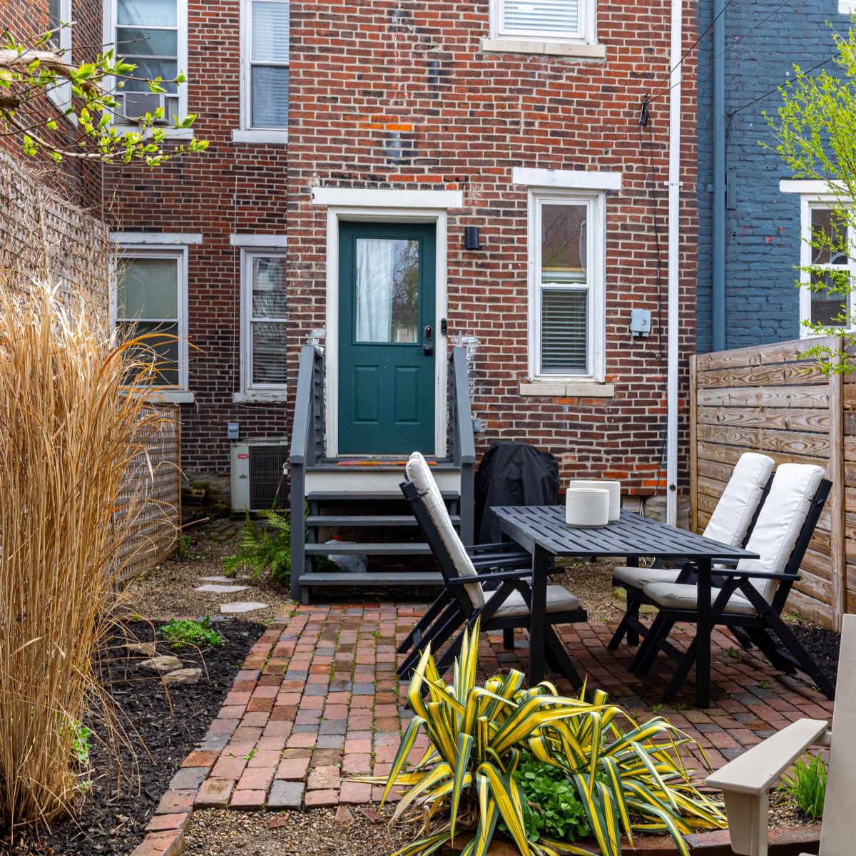 A small brick patio features a table and chairs, surrounded by plants and backyard structures, with a green door at the back of a red brick building.