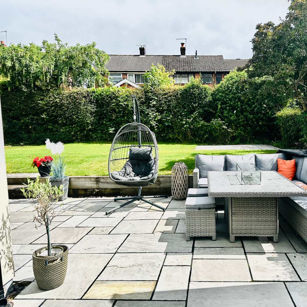 A landscaped backyard with a stone patio, a corner seating area with a glass table, and a hanging chair surrounded by greenery.