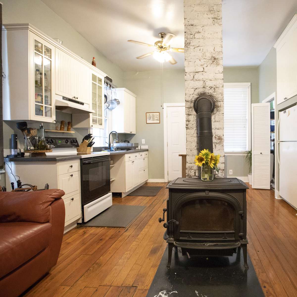 A kitchen with white cabinets, a stove, and a central farm-style table in a room featuring wooden floors and a brick chimney.