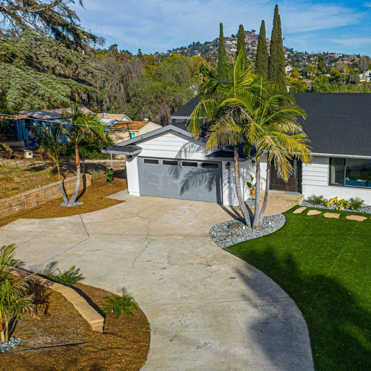 A modern single-family home with a two-car garage, a manicured lawn, and landscaping features including palm trees and a gravel path.