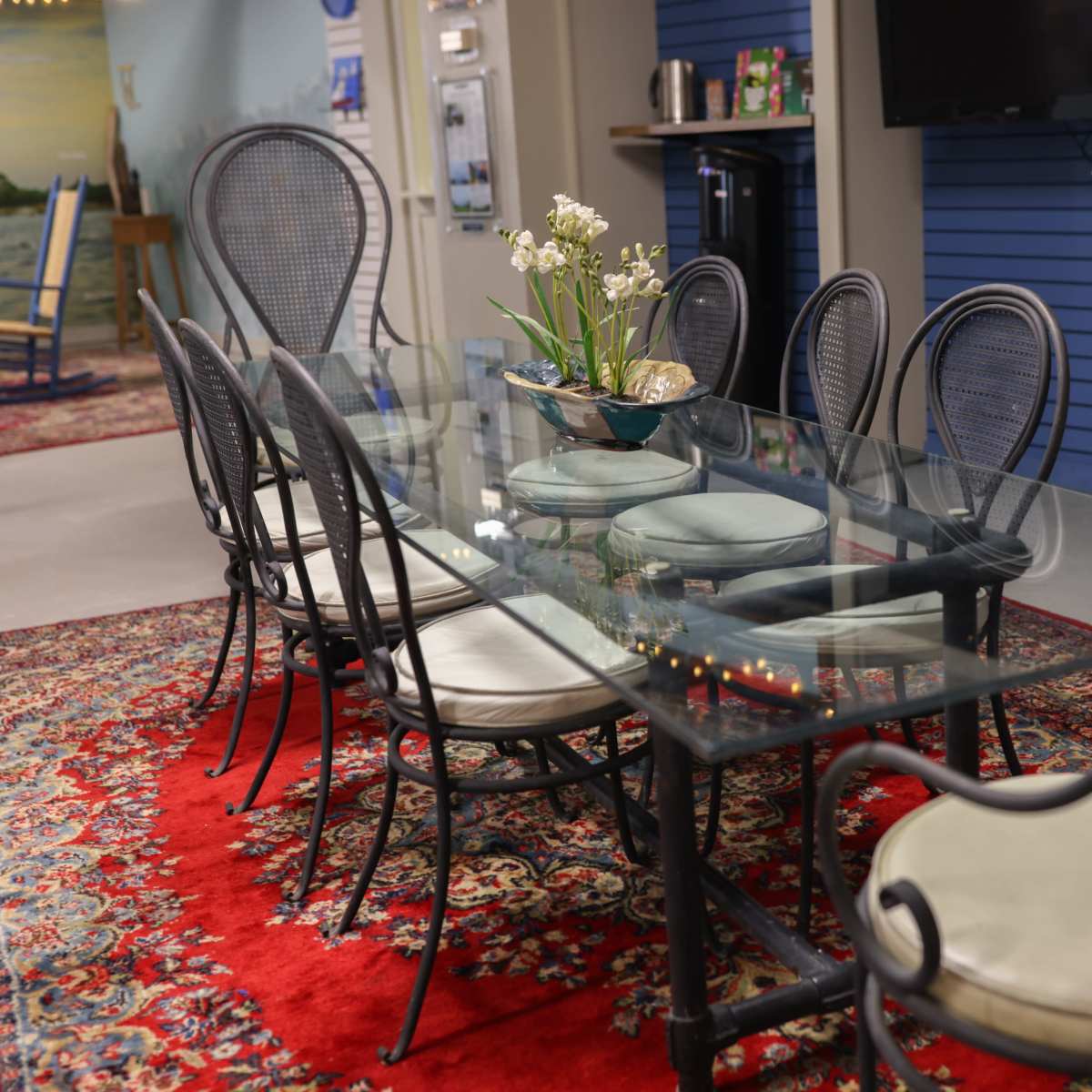 A glass dining table surrounded by eight chairs sits on a decorative red rug in a well-lit room.