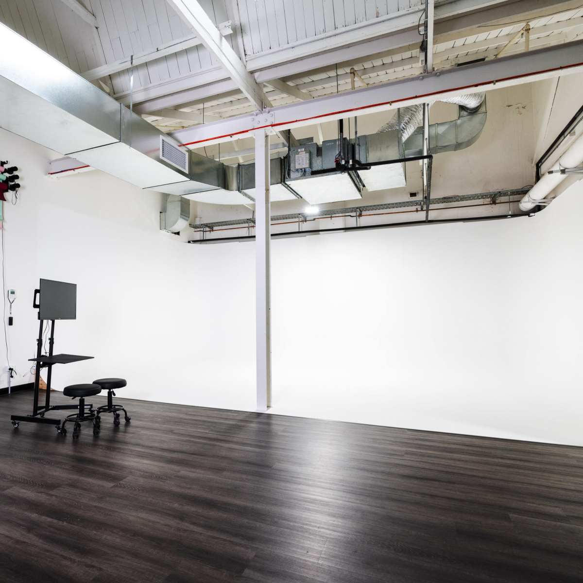 A spacious studio with a white backdrop, dark wooden floors, and a mobile monitor stand.
