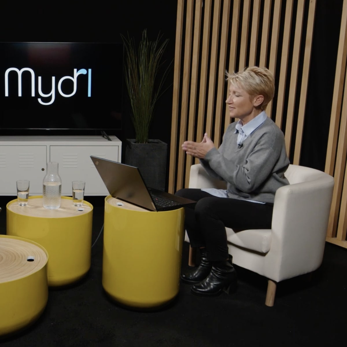 A man and a woman sit in modern chairs facing each other, with a laptop on a yellow coffee table between them, in a studio setting featuring plants and a television screen displaying the logo "Mydri."