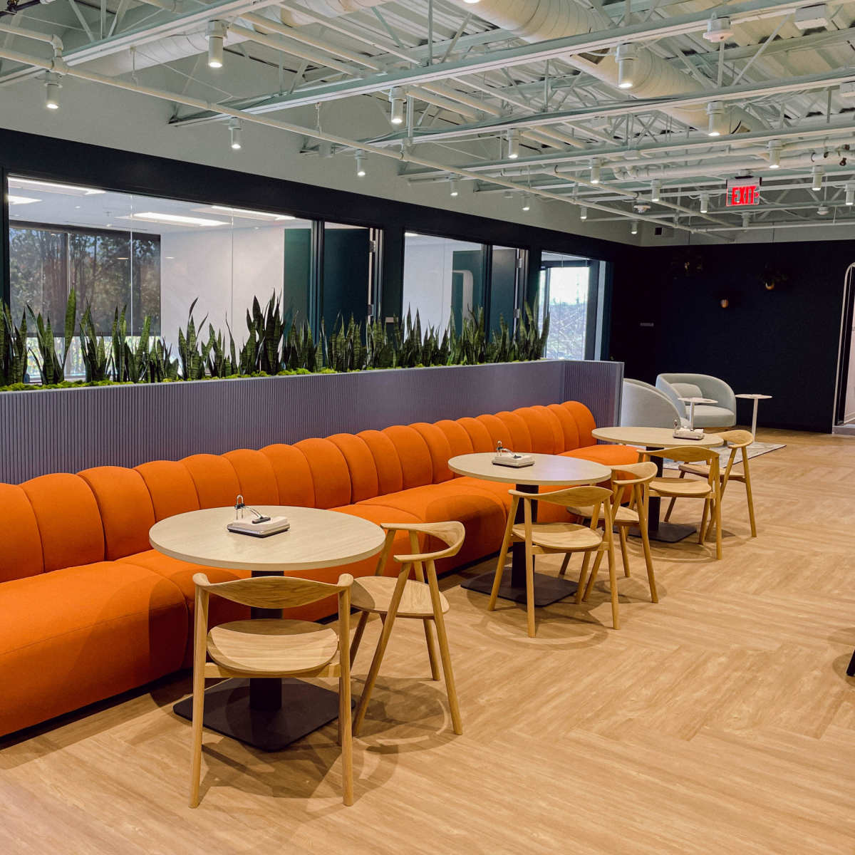 A modern lounge area featuring an orange curved couch and several round tables with wooden chairs, surrounded by indoor plants.
