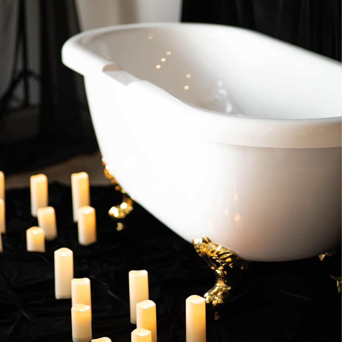 A white freestanding bathtub with gold feet is positioned on a dark surface surrounded by lit candles.