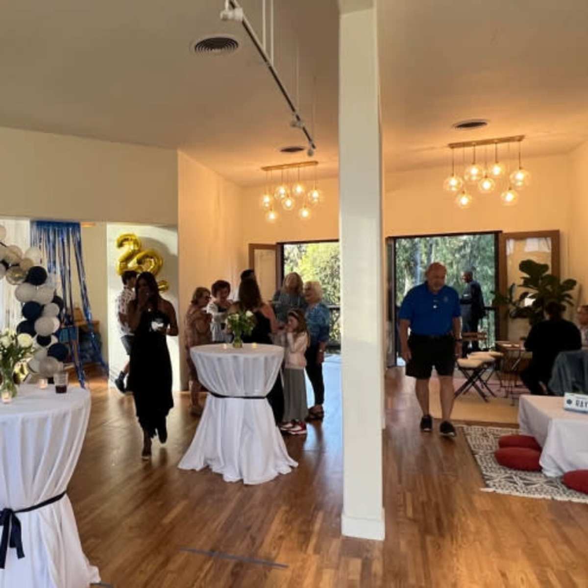 A group of people socializes at a spacious indoor event, featuring tables with refreshments and a decorative balloon arch in the background.