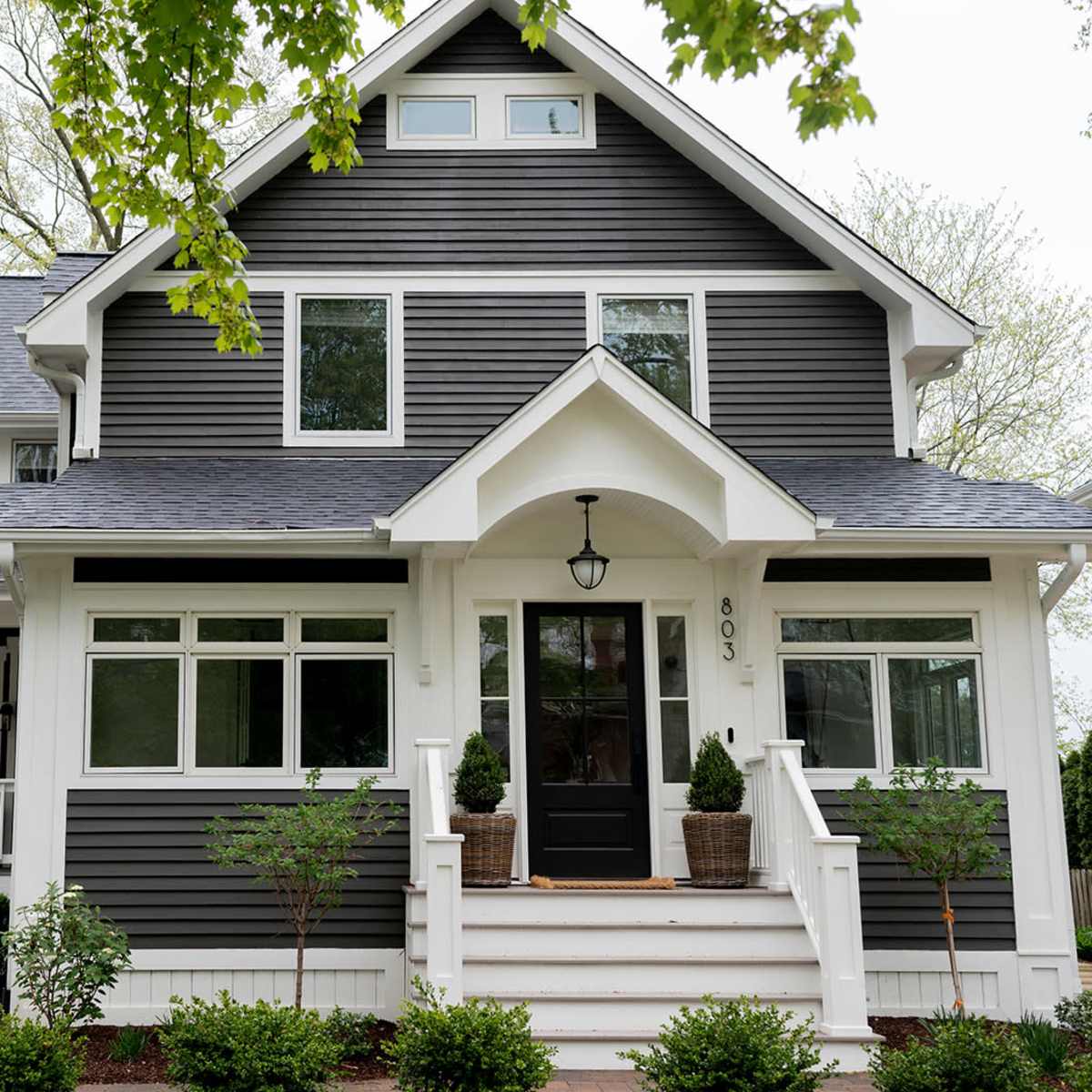 A large, two-story gray house with a steep roof, white trim, and a front porch flanked by neatly arranged shrubs.