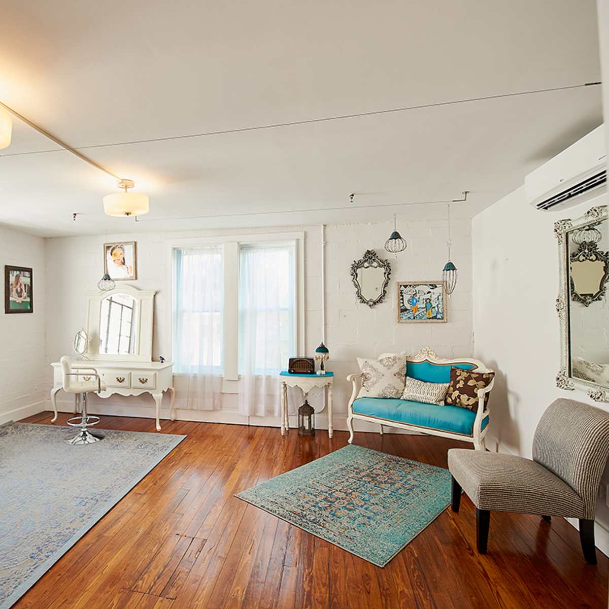 A bright, well-furnished room with wooden flooring, large windows, and various decorative mirrors and furniture pieces.