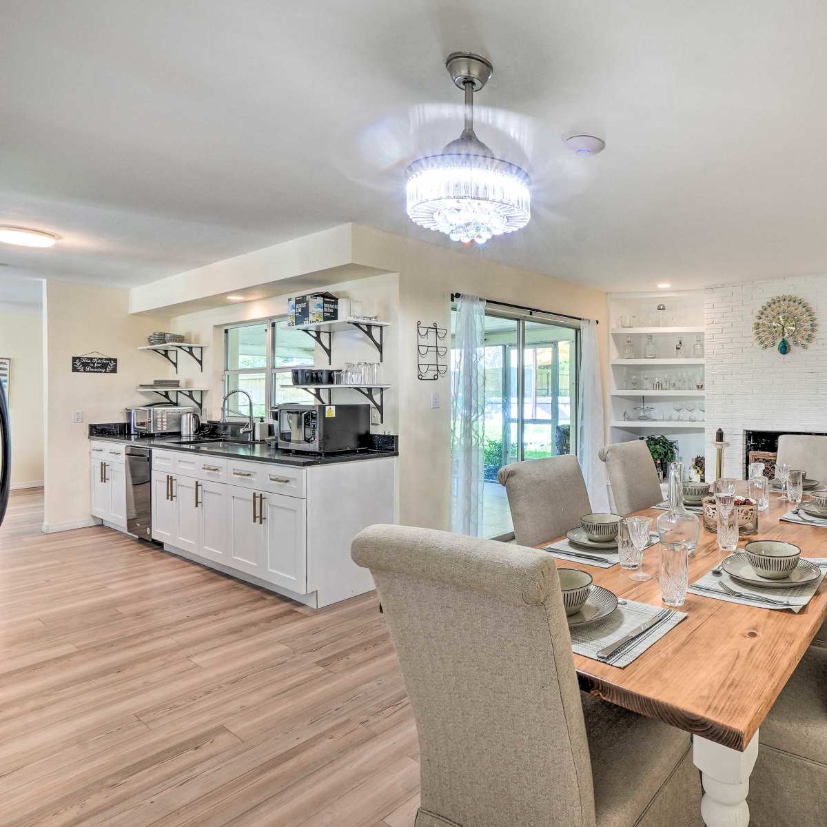 A dining room setup with a wooden table laid for a meal, surrounded by upholstered chairs, and a modern kitchen area with white cabinetry and open shelving in the background.