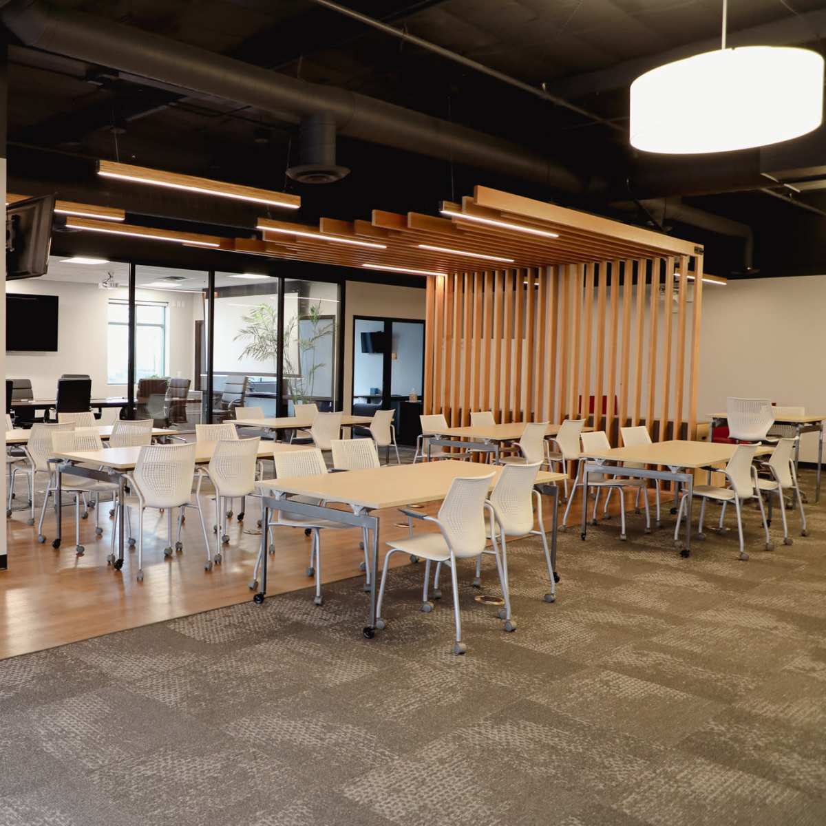 A modern office space with several tables and chairs arranged for collaborative work, along with glass-walled meeting rooms in the background.