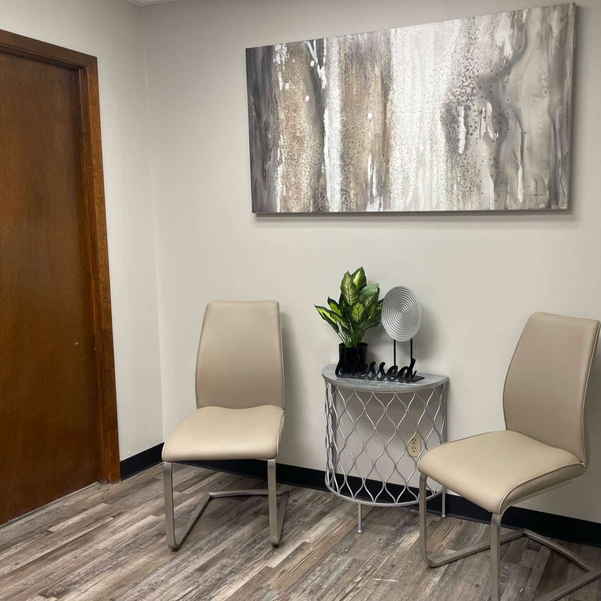 A minimalist waiting area with two beige chairs, a small round table, a decorative plant, and a fan against a neutral wall, featuring an abstract painting above.