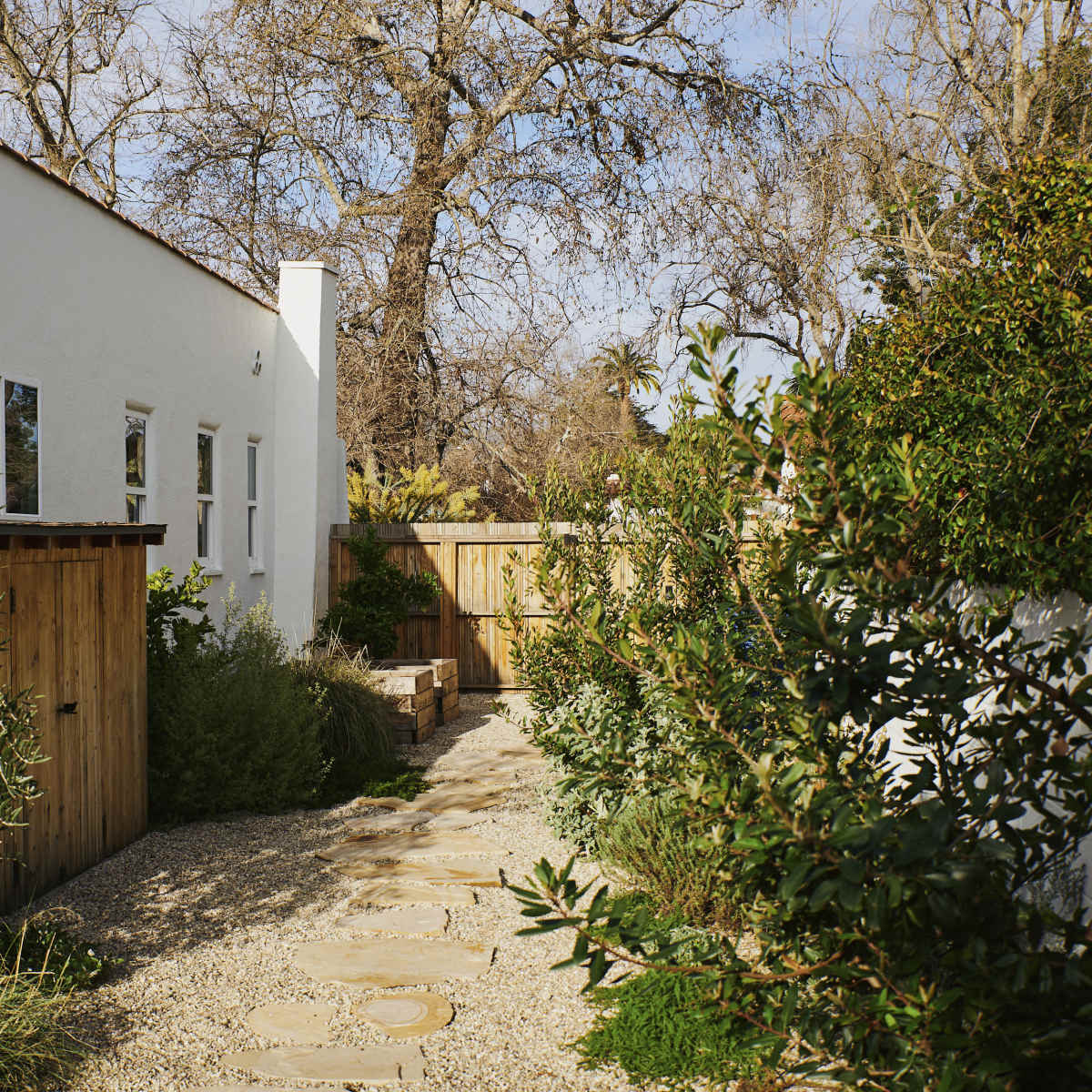 A stone path lined with greenery leads to a wooden gate beside a white building set in a tranquil outdoor space.