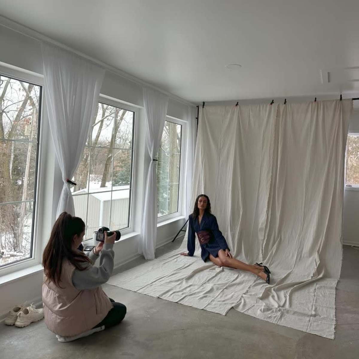 A photographer captures a model posing on a light-colored backdrop in a spacious room with large windows.