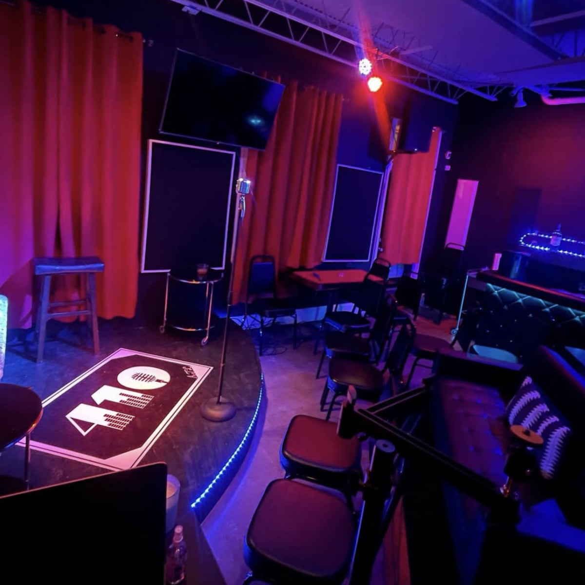 A dimly lit performance space featuring a stage with a microphone, illuminated seating areas, and red curtains.