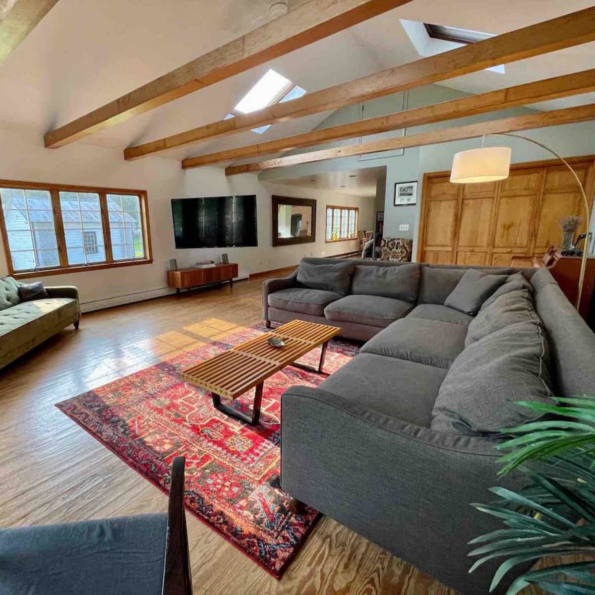 The living room features a sectional sofa, a green couch, a patterned rug, wooden beams, and a large window.