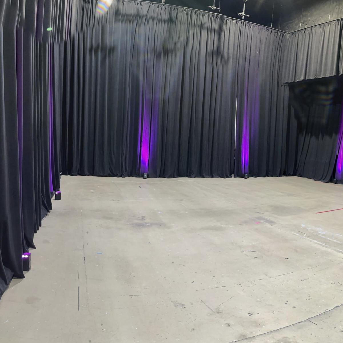 A large empty space with black curtains lining the walls and purple lights illuminating the floor.