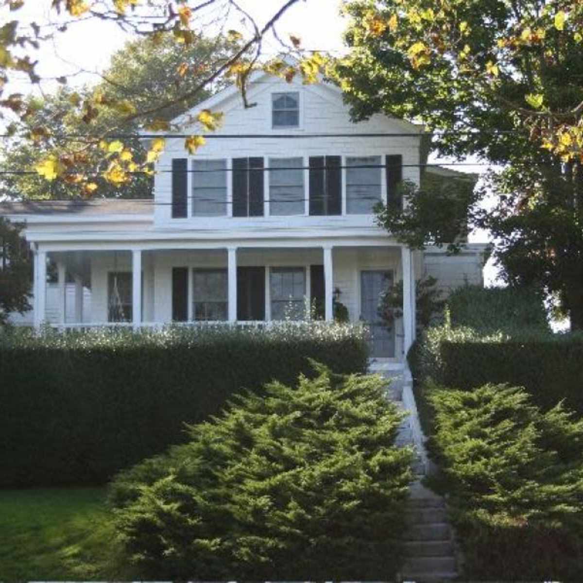 A white two-story house with black shutters is set behind a neatly trimmed hedge and a gravel pathway.