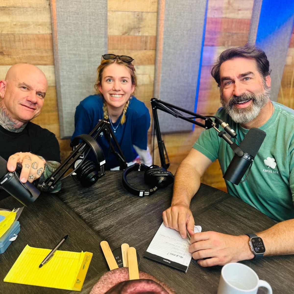 Three individuals sit at a table with microphones and recording equipment, surrounded by wooden paneling.