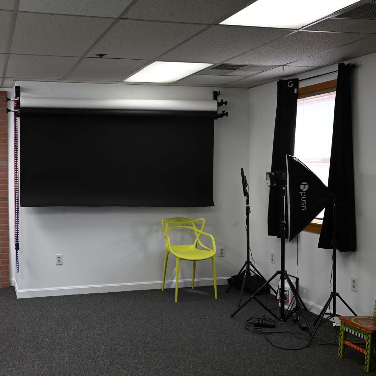 A photography studio with a black backdrop, two softbox lights, and a bright yellow chair against a brick wall.