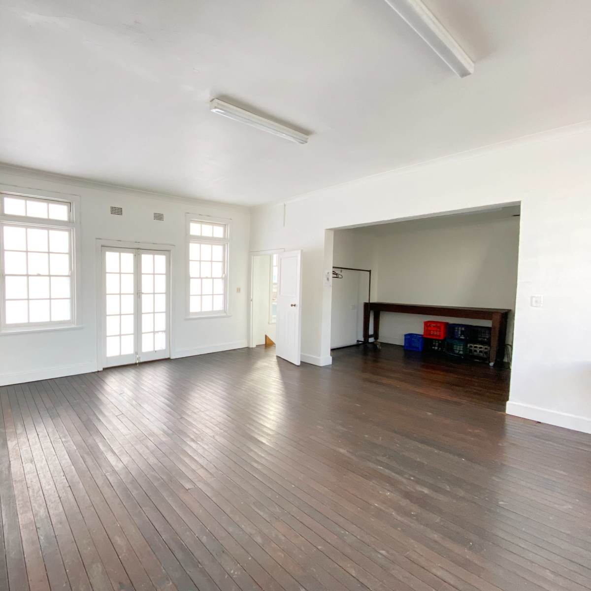 A spacious, empty room with wooden floors and large windows, featuring a table in the adjacent area.