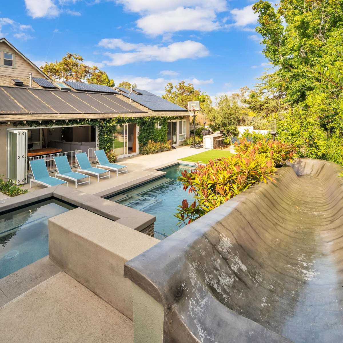 A modern backyard with a swimming pool, a water slide, and a house featuring solar panels and an outdoor seating area.
