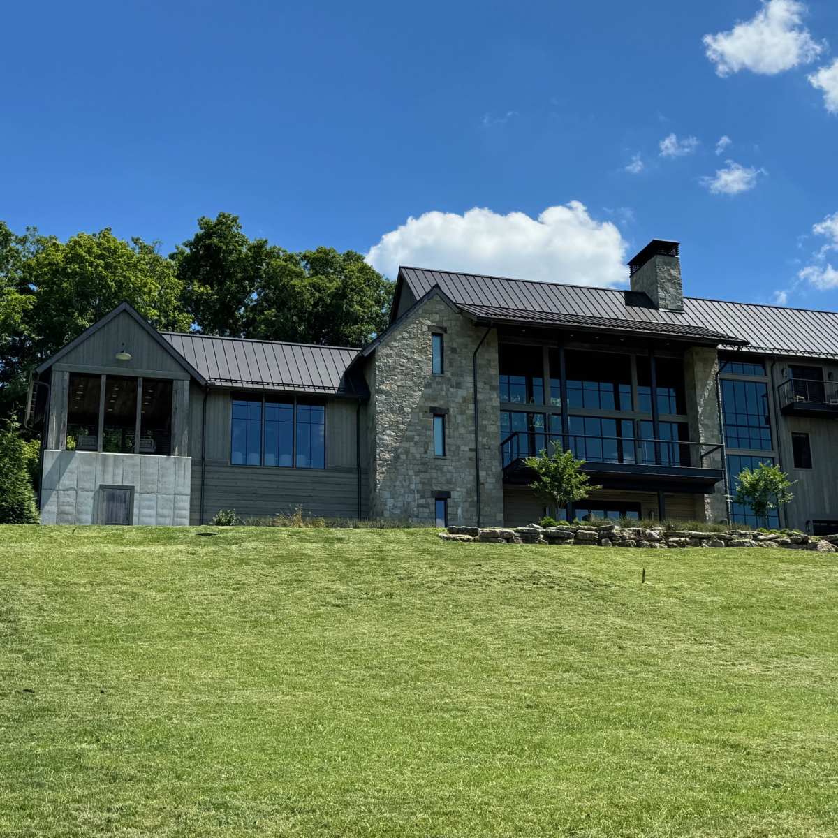 A modern stone and wood house with large windows, set on a grassy hillside under a clear blue sky.
