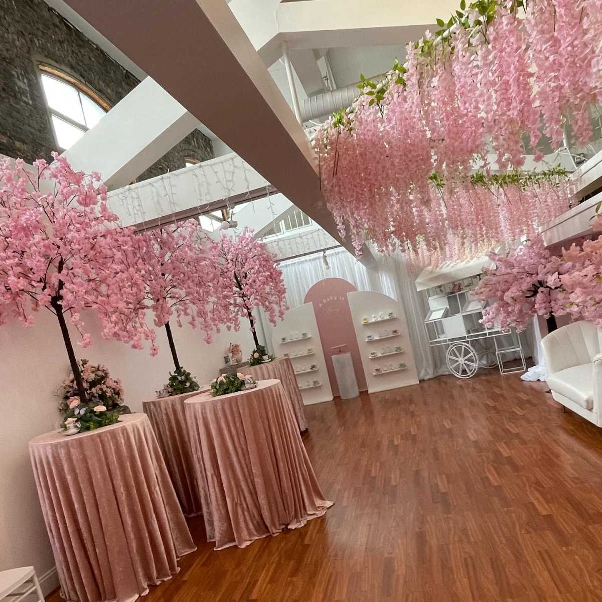 A spacious room decorated with pink wisteria trees, velvet-covered tables, and white decor elements, creating an elegant atmosphere.