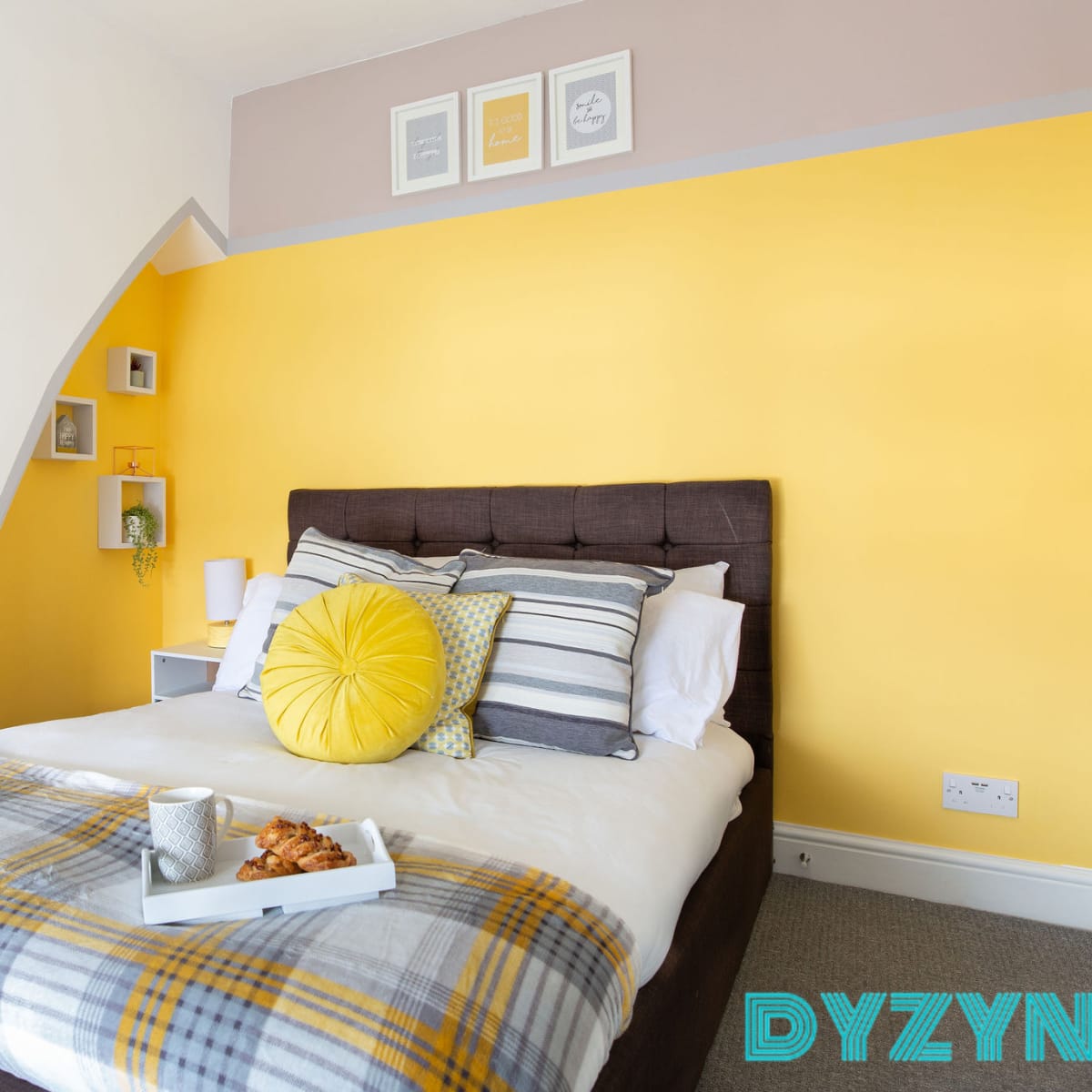 A brightly colored bedroom with a yellow accent wall, a neatly made bed featuring decorative pillows, and a tray on the bed with a cup and a snack.