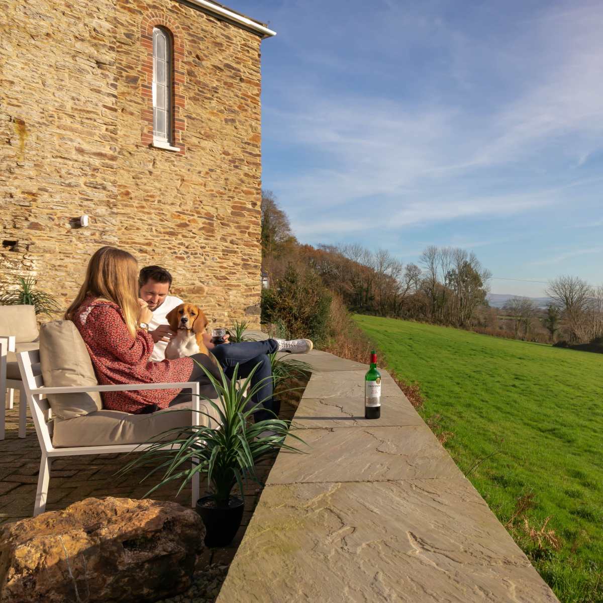A woman and a man sit on a stone terrace with a dog and a bottle of wine, overlooking a large green field and blue sky.