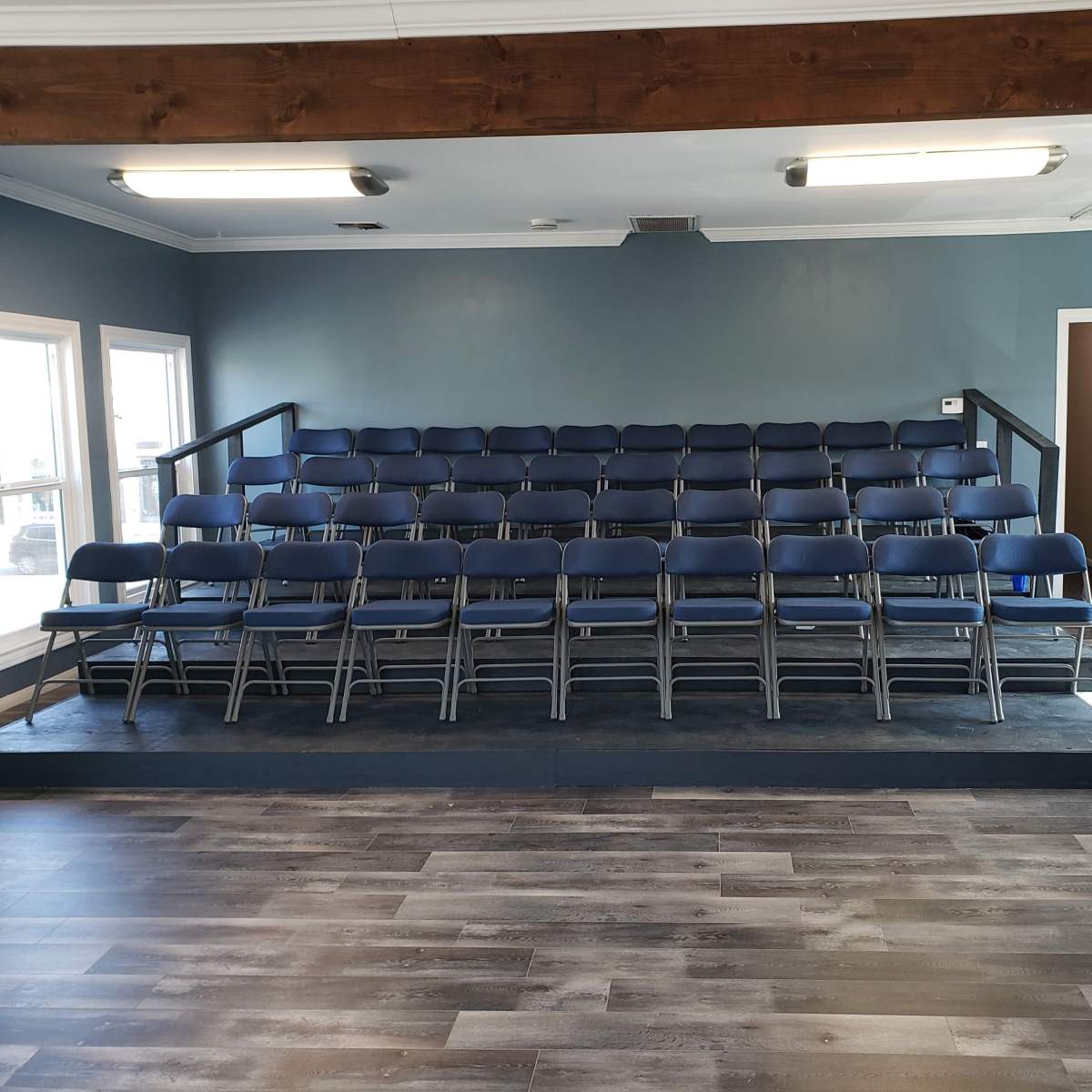 A small auditorium with stacked blue folding chairs arranged in a tiered seating layout and a wooden beam along the ceiling.