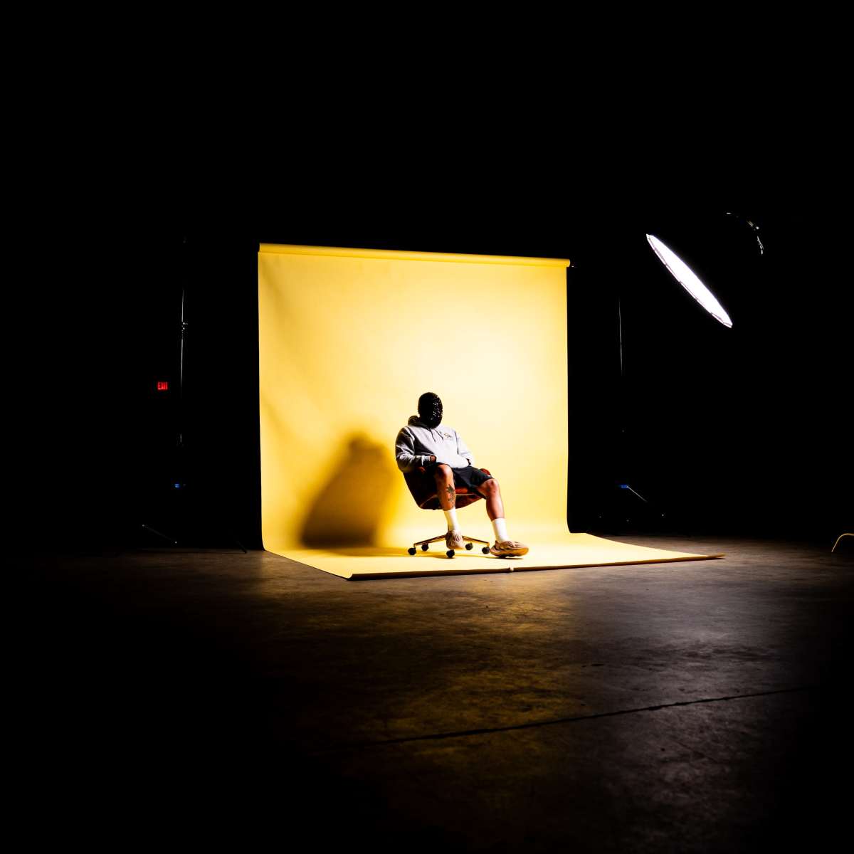 A person sits on a chair against a yellow backdrop in a dimly lit studio.