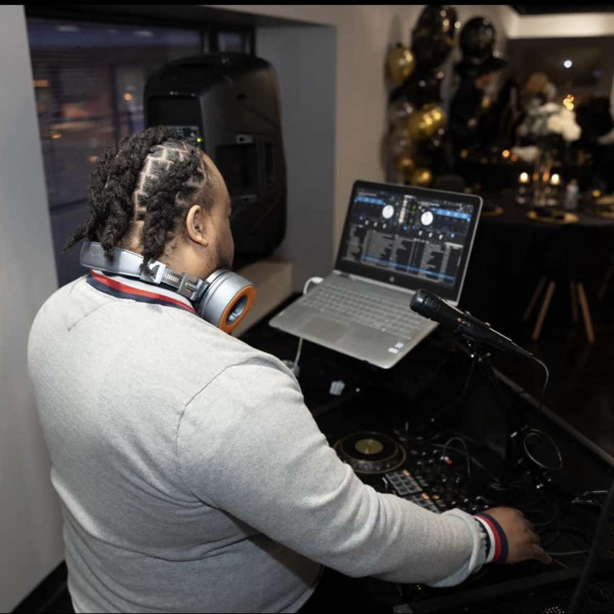A DJ stands at a setup with a laptop and mixing equipment, wearing headphones and facing away from the camera in a venue decorated with black and gold balloons.