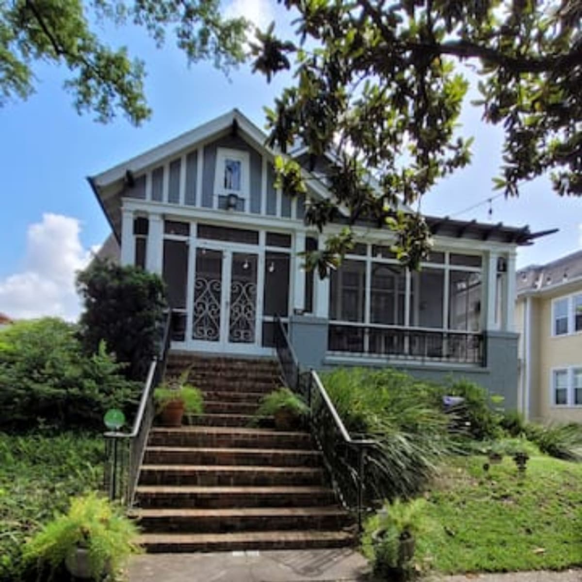 A two-story house with a front porch and decorative railings is set on a grassy yard with stone steps leading to the entrance.