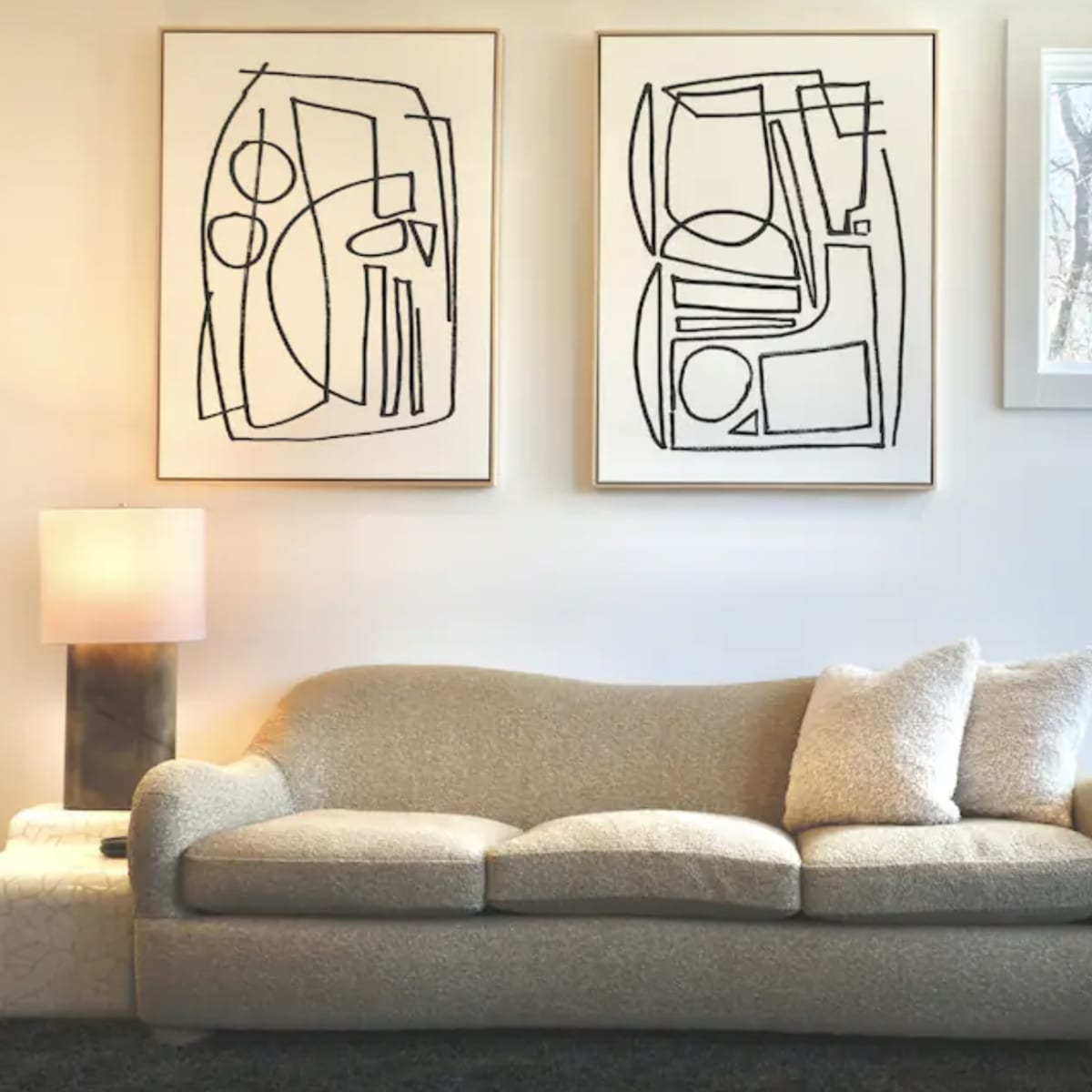 A living room features a light-colored sofa with textured upholstery, two abstract line art pieces on the wall, and a window allowing natural light to enter.