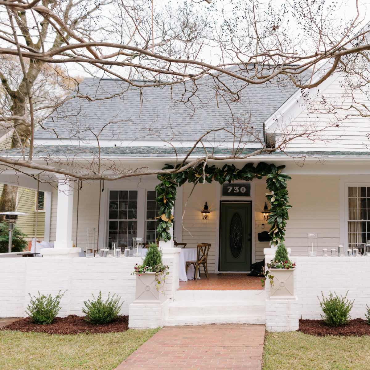 A white, single-story house with a covered porch is adorned with greenery around the entrance, featuring a brick pathway and neatly landscaped shrubs.