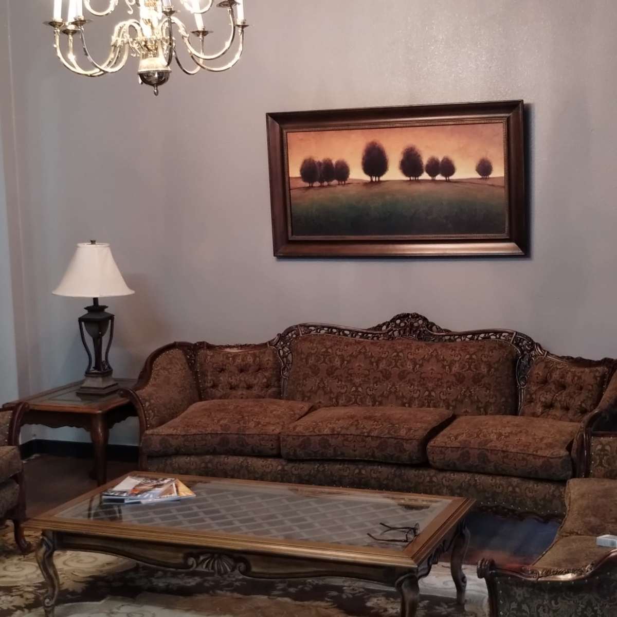 A living room furnished with a patterned couch and armchairs, a wooden coffee table, two lamps, and a chandelier, all set against a backdrop of green curtains and a framed landscape painting.