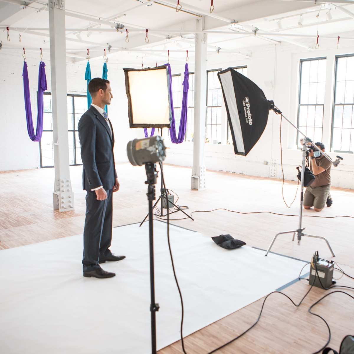 A man in a suit stands on a white backdrop, while a photographer kneels behind a camera and softbox in a well-lit studio.