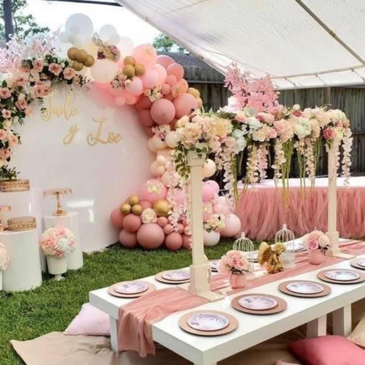A decorated outdoor party area featuring a floral backdrop with pink and gold balloons, a white table set with plates, and floral arrangements.