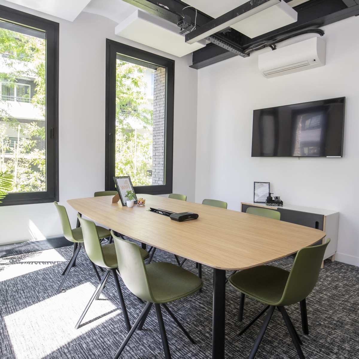 A meeting room features a large wooden table surrounded by green chairs, with a television mounted on the wall and large windows allowing natural light to enter.