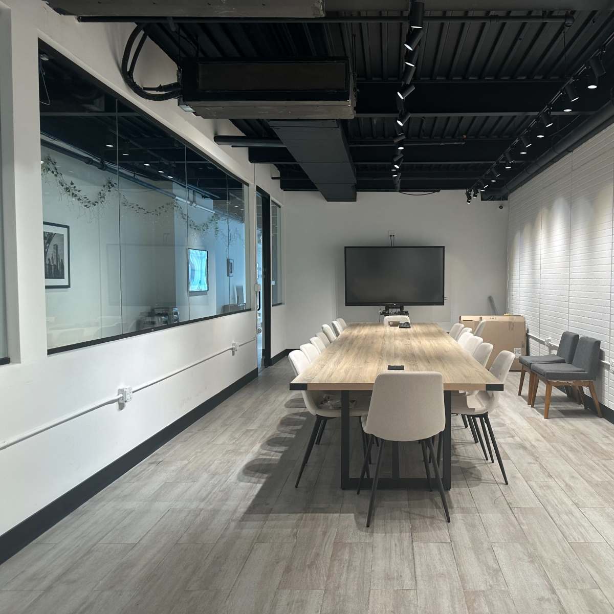 A long wooden table with chairs surrounds it in a well-lit conference room featuring a large TV on the wall.