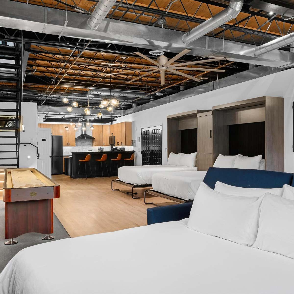 A modern open-concept living space featuring two beds, a kitchenette with seating, and a staircase leading to an upper level.
