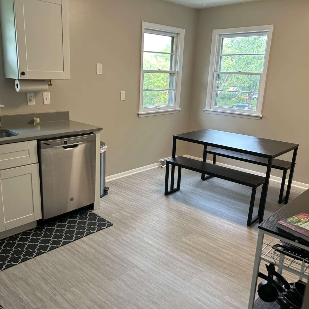 A modern kitchen features white cabinets, stainless steel appliances, and a small dining table beside two windows.