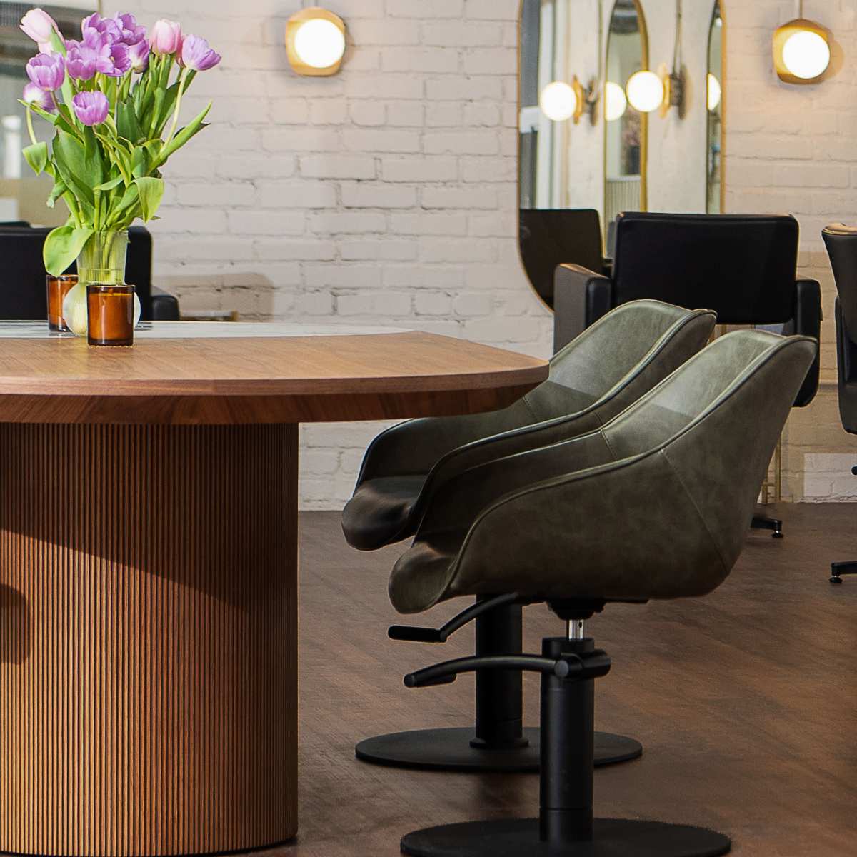 A modern salon interior features a round wooden table, green chairs, and a vase of purple tulips, accented by wall mirrors and soft lighting.