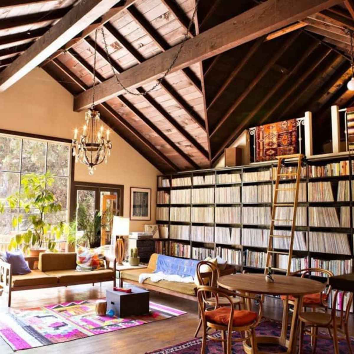 A spacious living room with high wooden ceilings, a large bookshelf filled with records, and comfortable seating area beside large windows.
