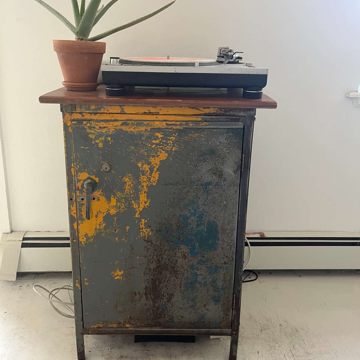 A vintage cabinet with a weathered finish holds a turntable on its top, while a potted aloe vera plant sits beside it.